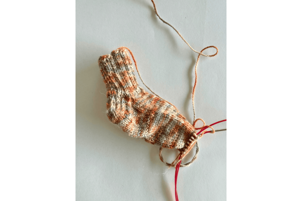 Hand-knit baby sock in orange, cream and grey alpaca blend, in progress on circular needles.